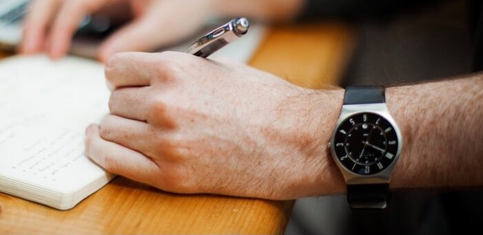 A hand holding a pen writes in a notebook, a watch is prominently featured.